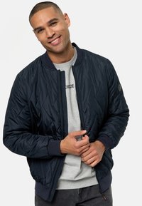 Chaqueta bomber acolchada de color navy con cremallera frontal, puños y cuello de canalé. Llevada sobre un sudadera gris de cuello redondo, con un sutil detalle del logo.