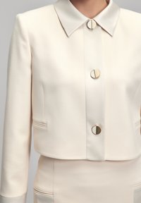 Krémový zkrácený blazer vyrobený z hladkého materiálu, s špičatým límcem, třemi zlatými kovovými knoflíky a detailním prošíváním na předních kapsách.