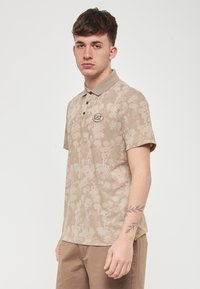 Polo beige con motivo floreale, maniche corte e colletto. Logo sul petto, apertura con cinque bottoni e tessuto testurizzato.