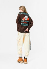 Sudadera de gran tamaño en marrón con estampado gráfico, pantalones cargo crema con bolsillo lateral y botas gruesas color marrón claro. Conjunto casual con un ajuste holgado.