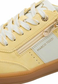 Zapatilla deportiva de ante amarillo con detalles en cuero blanco, que presenta una cremallera dorada y suela de goma texturizada. Incluye cordones blancos y detalles perforados.