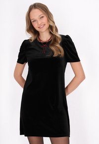 Rochie neagră din catifea, cu mâneci scurte tip balon, având decolteu rotund și un detaliu cu fundiță roșie la guler. Textură netedă, lungime deasupra genunchiului.