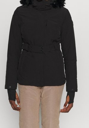 Schwarze Winterjacke mit Gürtel und mit Pelz besetzter Kapuze, getragen über beigen Hosen, mit Reißverschlusstaschen an Brust und Seiten sowie verlängerten Ärmelbündchen.