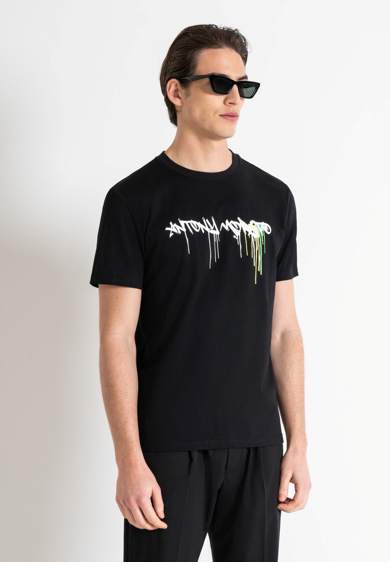 Camiseta negra de manga corta hecha de algodón con un estampado en blanco y verde de estilo graffiti. El modelo lleva gafas de sol negras y pantalones negros.