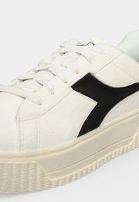 Baskets blanches avec semelle épaisse et logo angulaire noir sur le côté, avec des lacets blancs et une semelle texturée dessous.