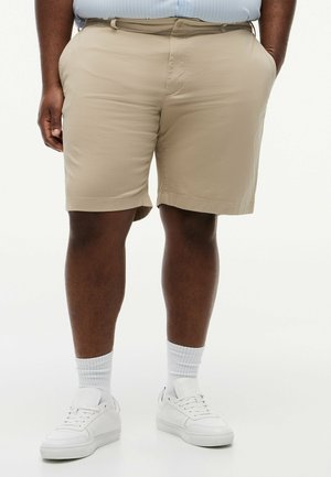 Mann trägt beige Shorts, weiße Sportsocken und weiße Sneakers, steht mit den Händen in den Taschen vor weißem Hintergrund.