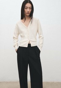 Cárdigan color crema con escote en v y cierre de botones, usado sobre pantalones negros de cintura alta. Material de punto suave con un ajuste relajado y mangas largas.