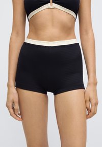 Svarte badeshorts med ribbemønster og kontrastfarget kremfarget midjebånd, med tettsittende design og glatt tekstur. Egnet for vannaktiviteter.