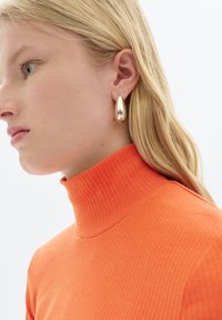 Goldene Tropfenohrringe mit reflektierender Oberfläche, getragen von einer Person mit einem orangefarbenen, gerippten Rollkragenpullover. Das schlichte, elegante Design ergänzt die Bekleidung.