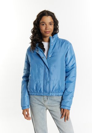 Ung kvinde med krøllet hår iført en lyseblå let dunjakke og lyseblå jeans mod en ensfarvet hvid baggrund.