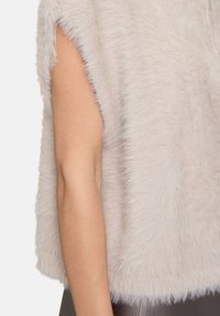 Nahaufnahme eines nackten Arms, der ein ärmelloses, hellbeiges, samtig strukturiertes Oberteil trägt, kombiniert mit einer Hose aus dunklem, glattem Stoff.