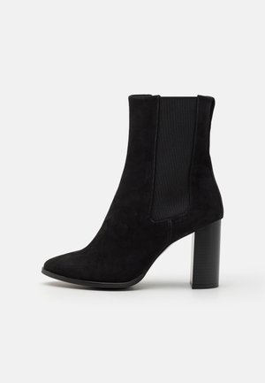 Bottine en daim noir avec un bout pointu, des panneaux latéraux élastiques et un talon épais noir. Texture lisse avec un design élégant et moderne.