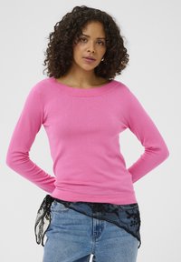 Mujer con cabello rizado que lleva una blusa de manga larga rosa brillante y unos jeans de mezclilla azul claro, con detalle de encaje negro en la cintura.