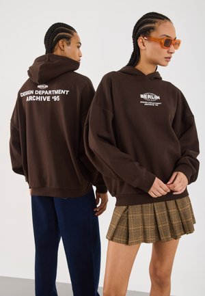 Brahno kapuco s puloverjem z belim besedilom na hrbtni strani, ki pravi "BERLIN DESIGN DEPARTMENT ARCHIVE '95." V kombinaciji s karirasto krilo.