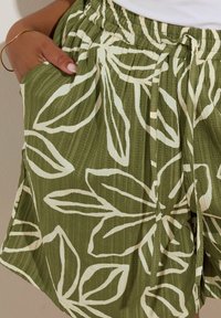 Pantaloni corti con vita elastica in tessuto verde oliva con una finitura texturizzata. Presentano grandi motivi a foglia bianca e tasche laterali.