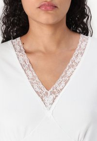 White garment with a V-neck featuring a delicate floral lace trim along the neckline. Smooth fabric with a soft texture.