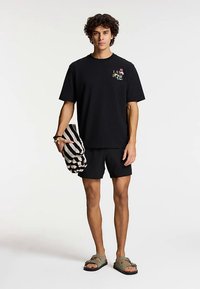 Černé bavlněné tričko s krátkými rukávy, zdobené barevným motivem. Kombinováno s černými šortkami a zelenými sandály, drží proužkovaný ručník.