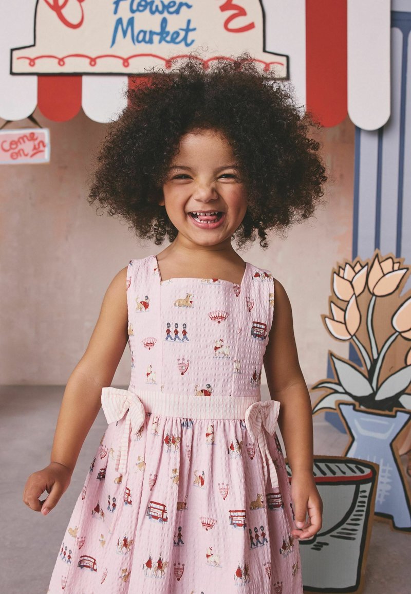 Niña sonriente con cabello rizado que lleva un vestido rosa con estampados temáticos de Londres, de pie frente a un fondo de mercado de flores con ilustraciones de tulipanes.