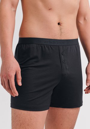 Männlicher Oberkörper und Oberschenkel, die schwarze Boxershorts mit geknöpftem Schlitz und gebrandeter Taille tragen, stehen vor einem neutralen Hintergrund.