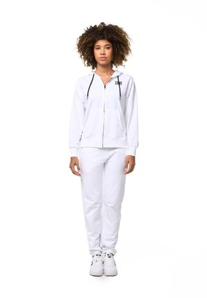 Un mannequin aux cheveux bouclés se tient devant un fond blanc, vêtu d’un sweat à capuche blanc zippé, d’un pantalon de jogging assorti et de baskets blanches.
