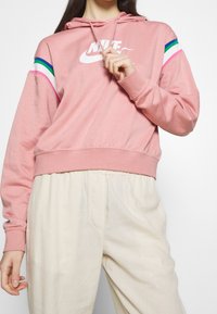 Rosa Nike-hoodie med vit logotyp, färgglada ränder på ärmarna, kort design, mjuk textur, matchad med ljusbeige byxor.