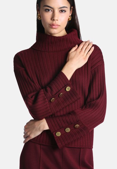 Femme portant un pull à col roulé côtelé bordeaux avec des boutons dorés sur les manches et de grandes boucles d’oreilles dorées, posant les bras croisés.