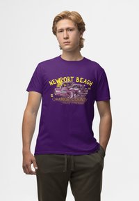 Fialové bavlněné tričko s grafikou klasického auta a textem "NEWPORT BEACH" a "ORANGE COUNTY SPEED SHOP." Krátké rukávy.