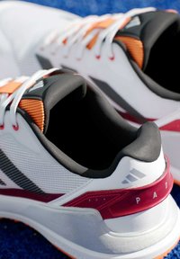 Witte sportieve schoenen met grijze en rode accenten, een textuur van mesh aan de bovenkant, een gepolsterde kraag en oranje details op de veters en buitenzool.