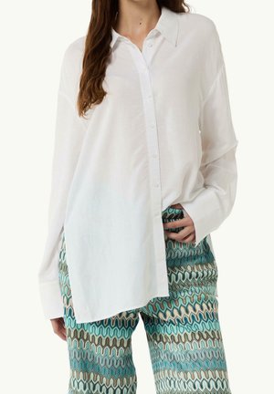 Kvinde iført en løs, hvid skjorte med lange ærmer og mønstrede bukser med blå, beige og sorte bølgemønstre, hånd hviler i lommen.