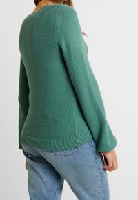 Grönstickad tröja med rund halsringning, ribbade ärmslut och kant, med en avkopplad passform och strukturerad stickad mönster. I kombination med ljusblå jeans.