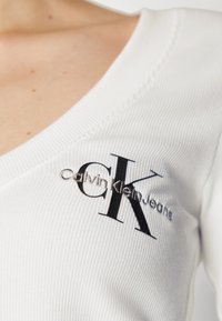 Vit ribbad kortärmad topp med V-halsning och en svart broderad logo "CK" tillsammans med "Calvin Klein Jeans" på vänster sida.