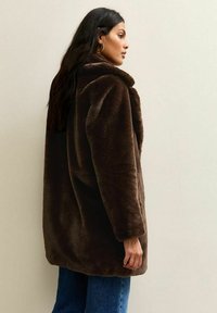 Bruine nepbontjas met een brede kraag en verlaagde schouders. Zachte textuur, middel-lang ontwerp gecombineerd met blauwe spijkerbroeken.