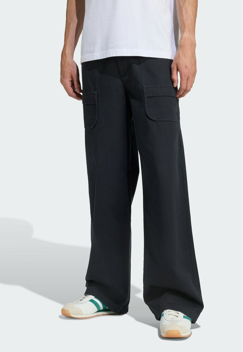 Man met wijde zwarte cargo broek met klepzakken en witte sneakers met groene strepen, staand tegen een effen achtergrond.