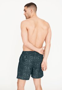 Tmavě vzorované plavkové šortky s elastickým pasem. Obsahují jemný květinový design s kontrastní texturou. Model je otočen zády k fotoaparátu.