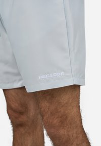 Lys blå shorts laget av glatt stoff, med en brodert "PEGADOR"-logo i hvitt, med rett snitt og sidesømmer.