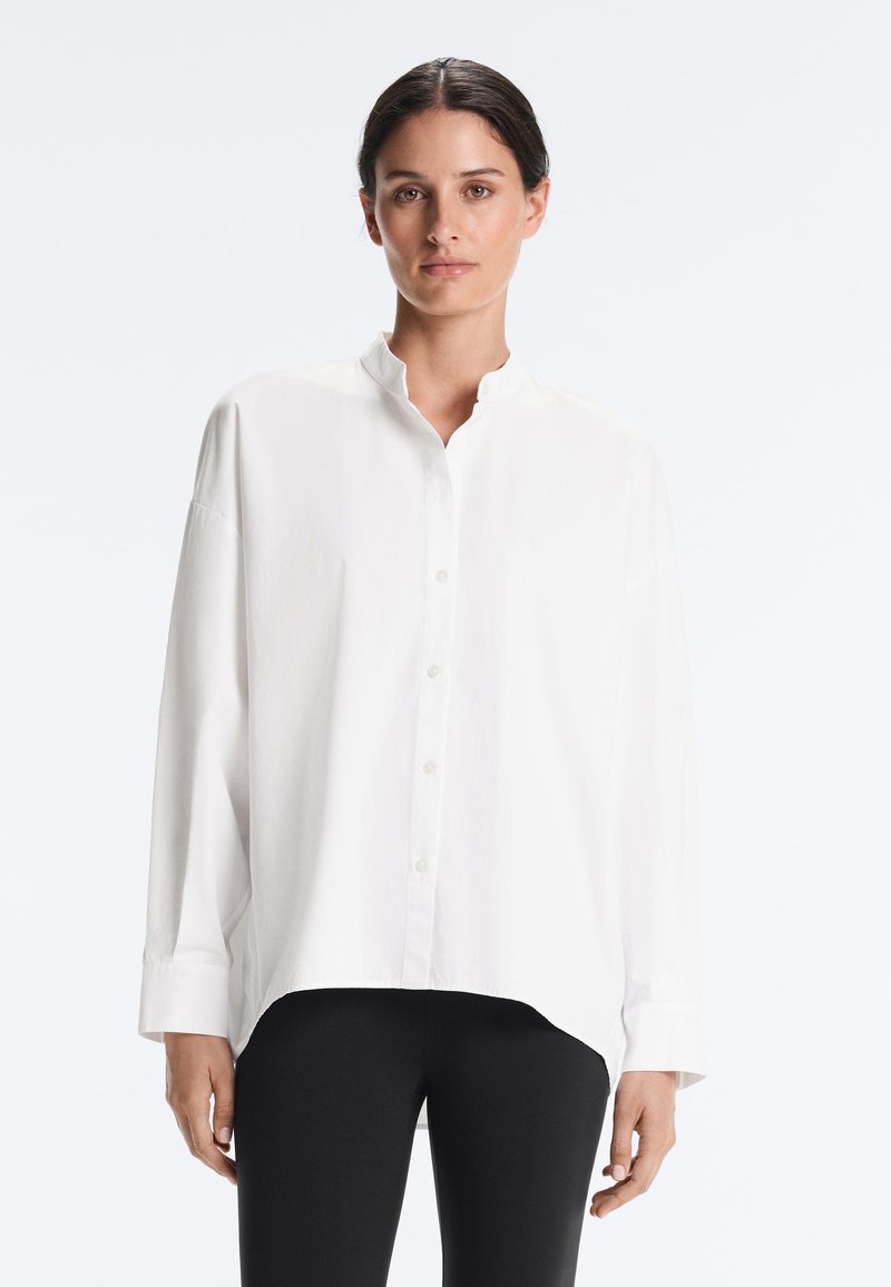 Femme aux cheveux foncés portant une chemise blanche ample boutonnée et un pantalon noir, debout devant un fond clair uni.