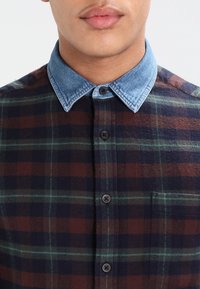 Geruit shirt met een blauwe denim kraag, bruine, marineblauwe en groene vierkanten, en een borstzak. Gemaakt van een zachte, gestructureerde stof.