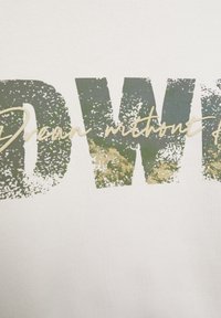 Witte trui met grote, verweerde groene letters die "Droom zonder angst" in een sierlijk lettertype weergeven. Zachte stofstructuur.