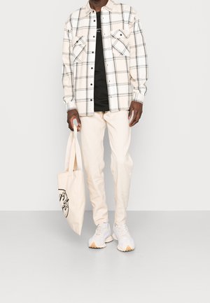 Camicia a quadri beige con dettagli neri, maglia nera sotto, pantaloni bianchi e sneakers chiare. La borsa presenta un design grafico minimalista.