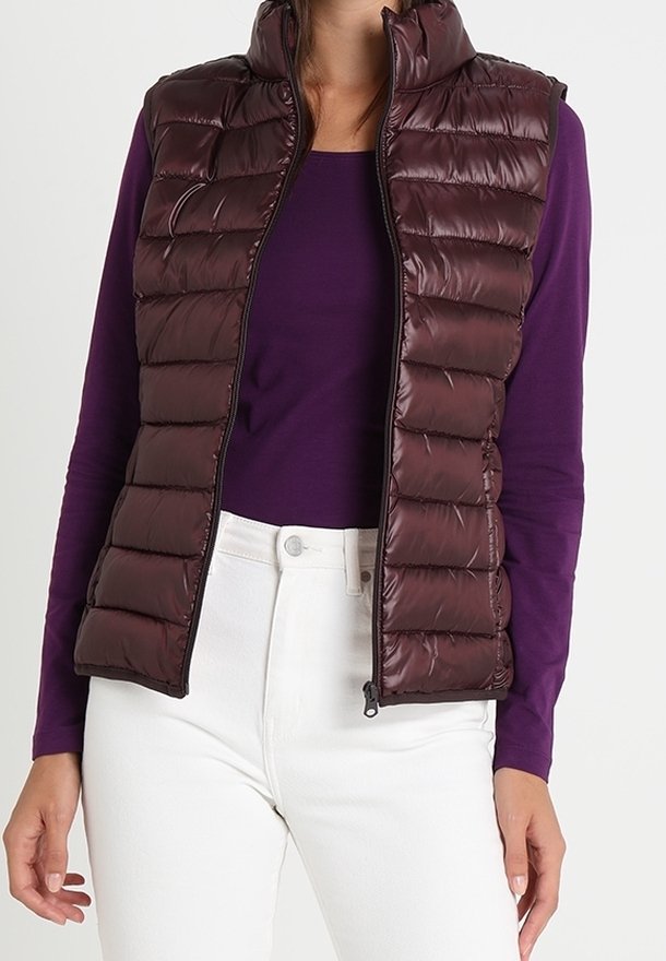 Femme portant une veste matelassée sans manches violet foncé sur une chemise violette à manches longues et un pantalon blanc, debout devant un fond uni.