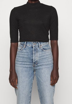 Sort glitrende tætsiddende top med albuelange ærmer kombineret med lyseblå, højtaljede denimjeans på en person med mørk hud.