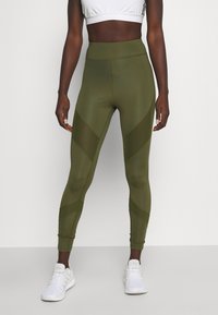 Hoge taille olijfgroene leggings met diagonale meshpanelen, gladde textuur en een aansluitend ontwerp, gecombineerd met witte sportschoenen.