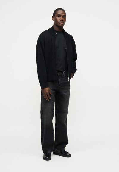 Νεαρός άνδρας που στέκεται, φορώντας μαύρο φούτερ με φερμουάρ, μαύρο t-shirt, ξεθωριασμένο μαύρο τζιν και μαύρα παπούτσια, μπροστά σε απλό λευκό φόντο.