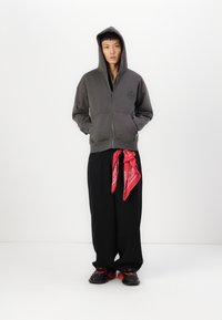 Sweat à capuche gris zippé avec un logo sur la poitrine, associé à un pantalon noir ample et un bandana rouge noué à la taille. Des chaussures noires complètent le look.