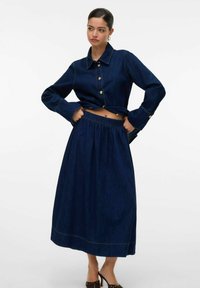 Denim kombinacija, ki vključuje temno modro skrajšano jakno z zlatimi gumbi in usklajen dolgo krilo z elastiko v pasu in šivom na robu.