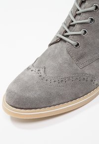 Chaussure en daim gris à bout arrondi, ornée de perforations décoratives et de lacets gris. La semelle est beige avec une bordure blanche sur le pourtour.
