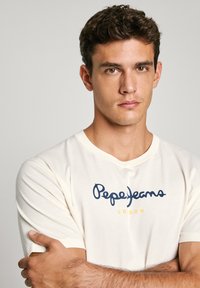 Krēmskrāsas T-krekls, kas izgatavots no kokvilnas, ar apaļu dekoltē. Ir tumši zilā "Pepe Jeans" logotips un dzeltenie akcenti. Īsas piedurknes.