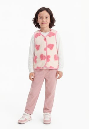 Enfant debout portant une chemise blanche à manches longues, un gilet sans manches rose et blanc avec des motifs de cœur, un pantalon en velours côtelé rose et des baskets roses.