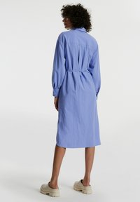 Hellblaues Hemdkleid mit Kragen, langen Ärmeln, tailliertem Schnitt und Rückenschlitzdetails. Getragen mit klobigen beigen Schuhen. Glatte Stofftextur.