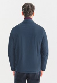 Granatowa kurtka o miękkiej teksturze, z podniesionym kołnierzem i bez widocznych elementów metalowych. Charakteryzuje się prostym designem i lekko dopasowanym krojem.